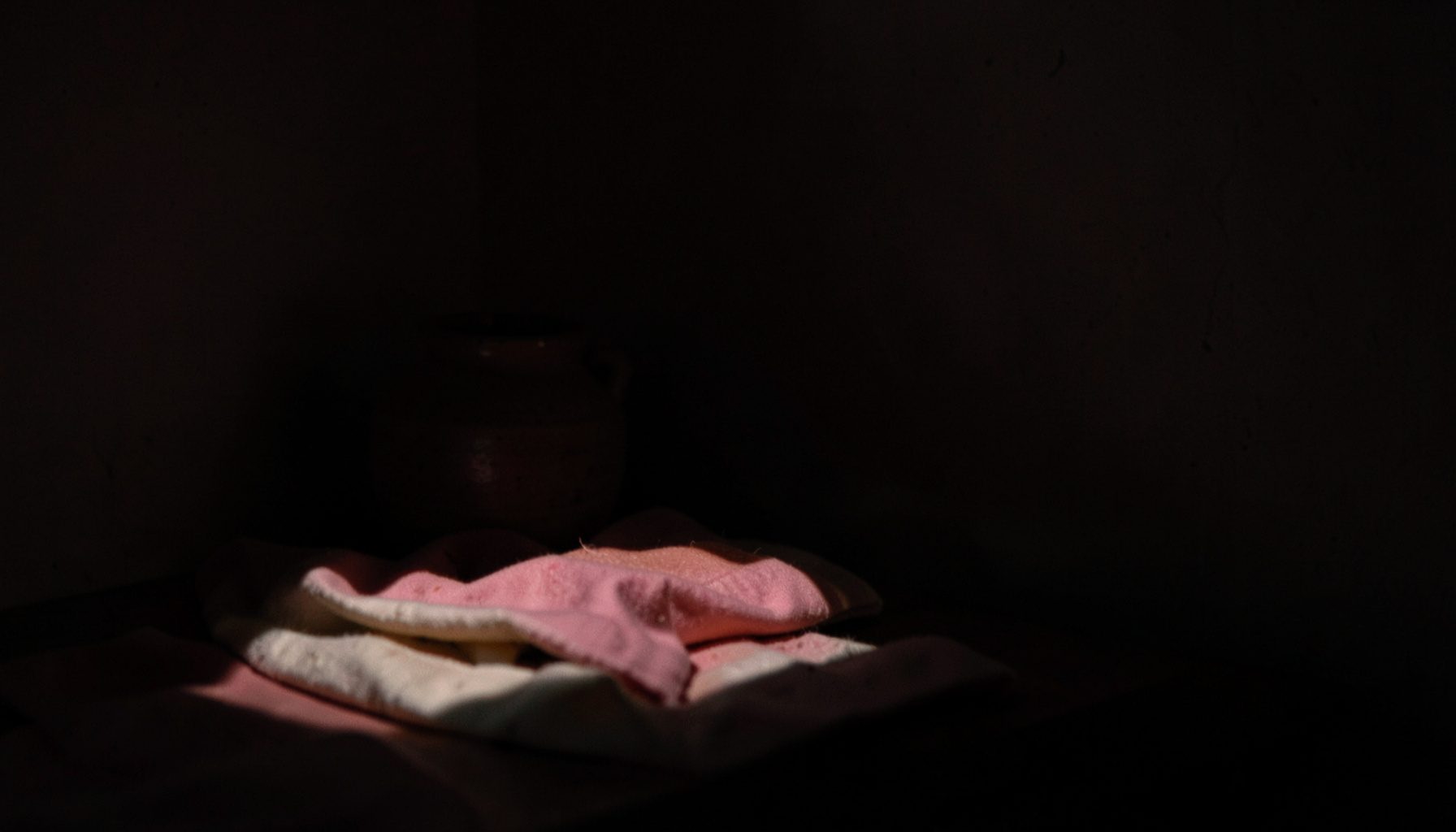 A dim workspace corner with a small ceramic vessel and folded fabric in dusty pink and cream tones