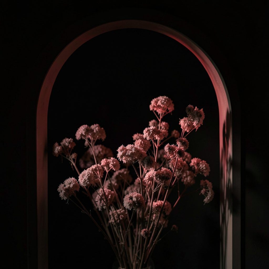 A dark moody arched frame view of dried flowers in dusty rose tones against deep shadow