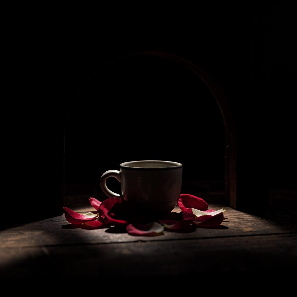 A shadowed arched frame still life of a small ceramic cup and rose petals on dark wood