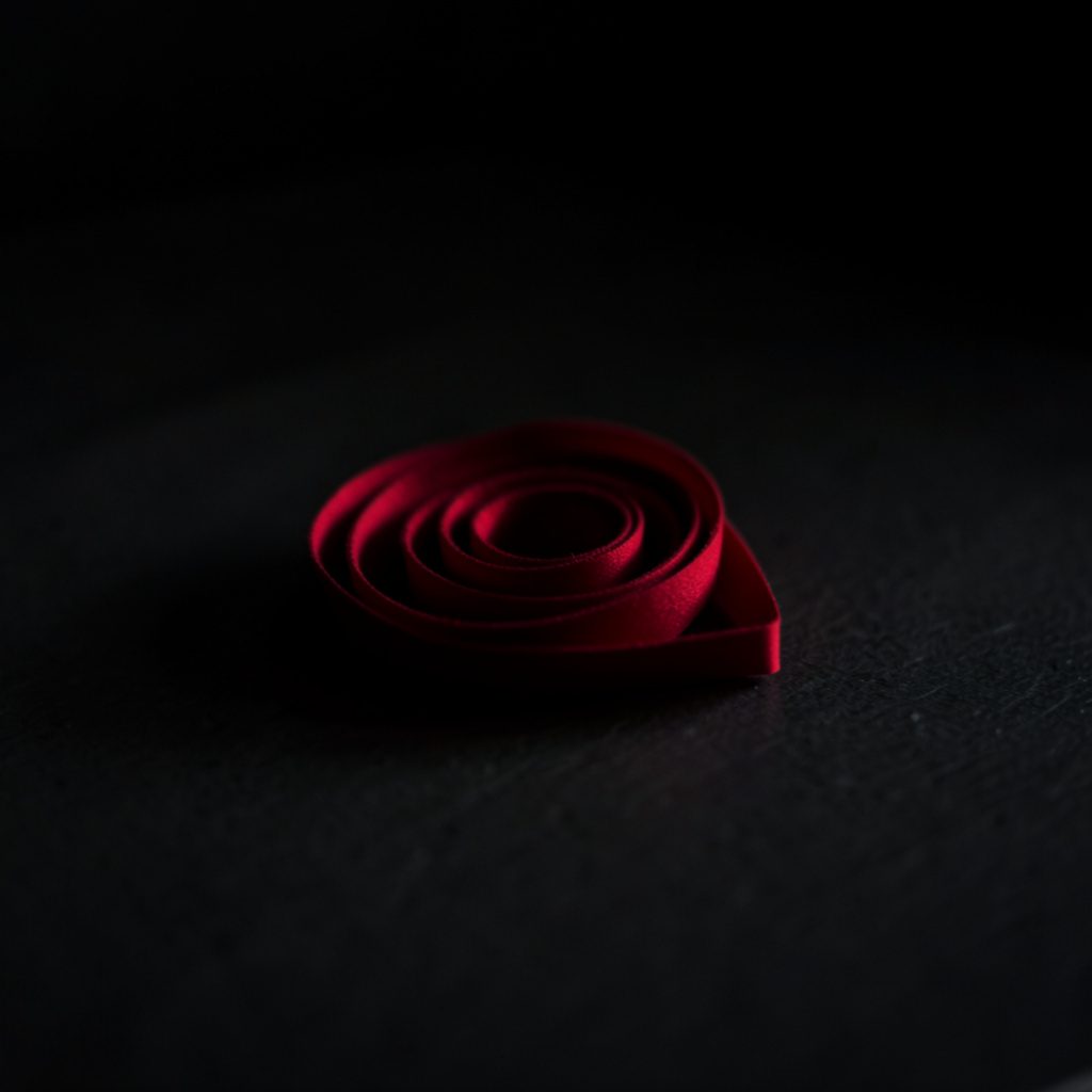 A small dark square photo of a rose colored ribbon coiled on a shadowed table